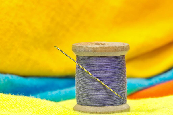 Carrete de hilo de algodón morado y aguja de coser sentado en la ropa de color brillante. Concepto de sastrería textil con enfoque en primer plano. - Foto, Imagen