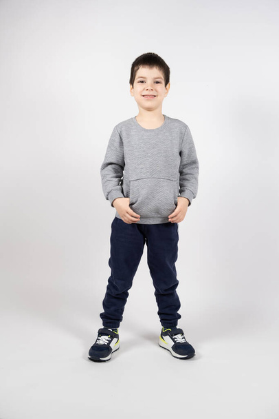 Ein kleiner Junge in Sweatshirt, Jogginghose und Turnschuhen auf weißem Hintergrund. Kinderbekleidung und Kinderschuhe. - Foto, Bild