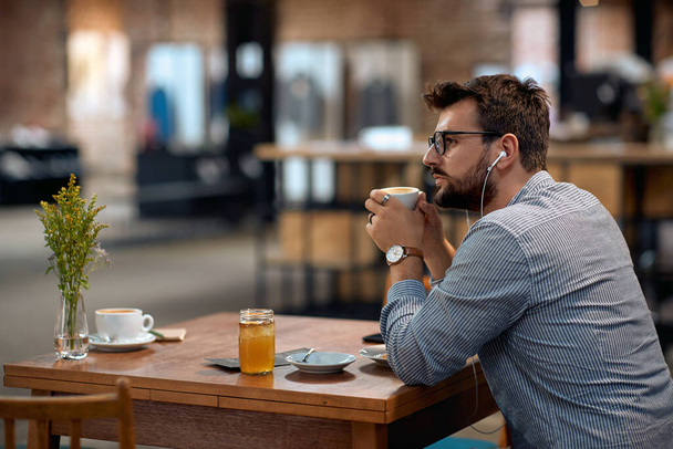 Man with earphones enjoying cup of coffee at cafeteria, looking into distance, pondering something. Lifestyle, lesiure concept. - Foto, Bild