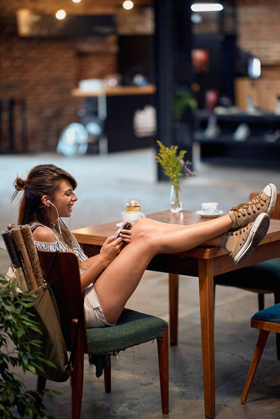 Beautiful young hipster girl with earphones, sitting alone at table with legs up feeling joyful in cafe. Lifestyle, student concept. - Photo, image