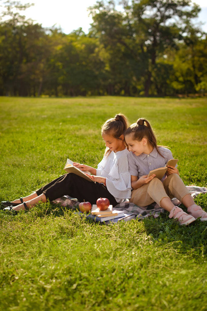 Niños positivos niñas sentadas en cuadros sobre hierba, leyendo libros, comiendo frutas. dos amigos despreocupados haciendo la tarea después de clases - Foto, imagen