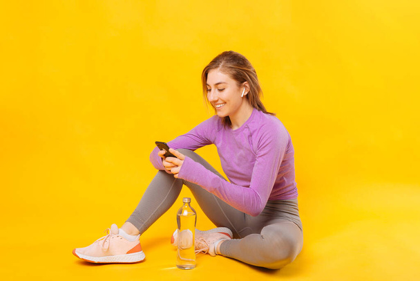 Veselá žena si dává pauzu od cvičení s telefonem a sluchátky. Studio záběr přes žluté pozadí. - Fotografie, Obrázek