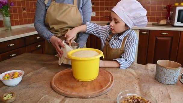 Heerlijk kindmeisje 5-6 jaar oud, bakkerssnoepje in witte koksmuts en beige schort, met een houten spooon, bloem met deeg mengen tijdens het koken van paastaart of panetonne thuis keuken - Video