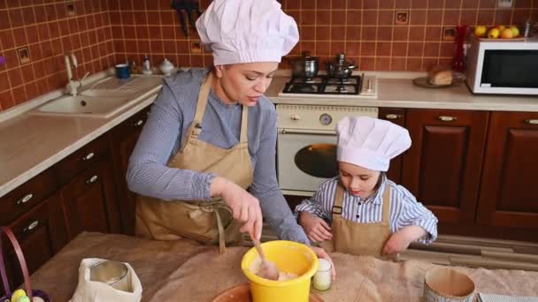 Gelukkige multi-etnische vrouw van middelbare leeftijd, een liefhebbende moeder en haar schattige dochter, schattig kind koken samen een kleine paastaart voor de paastafel in het gezellige houten keukeninterieur - Video