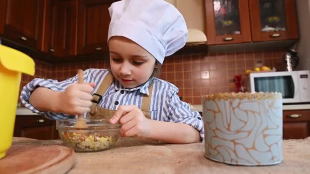 Белая 5-летняя девочка в белой шляпе шеф-повара и бежевом фартуке, с деревянной ложкой, смешивая сахар с изюмом в стеклянной чаше. Очаровательный ребенок учится готовить на домашней кухне. Кулинария - Кадры, видео