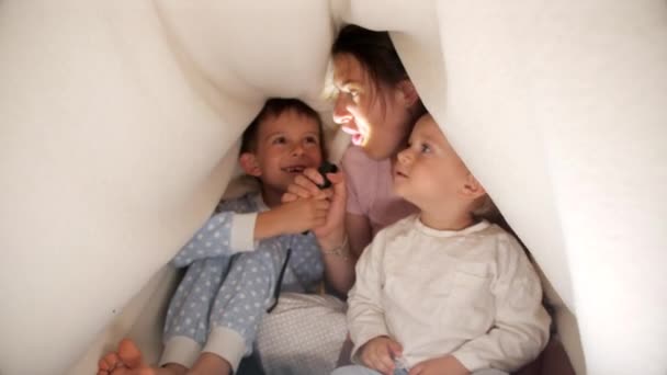 İki neşeli çocuk battaniyenin altında genç anneyle eğleniyor ve el feneriyle oynuyorlar. Ailenin birlikte vakit geçirmesi, ebeveynlik, mutlu bir çocukluk ve eğlence. - Video, Çekim