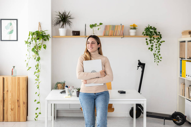 Уверенная предпринимательница, держащая в руках ноутбук, стоящий в домашнем офисе. Портрет счастливой хозяйки на рабочем месте - Фото, изображение