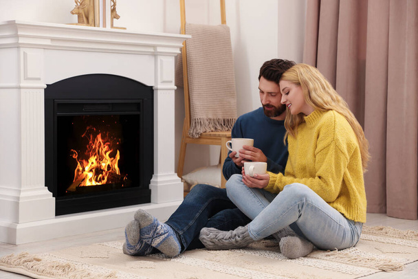 Lovely couple with hot drinks spending time together near fireplace at home - Φωτογραφία, εικόνα