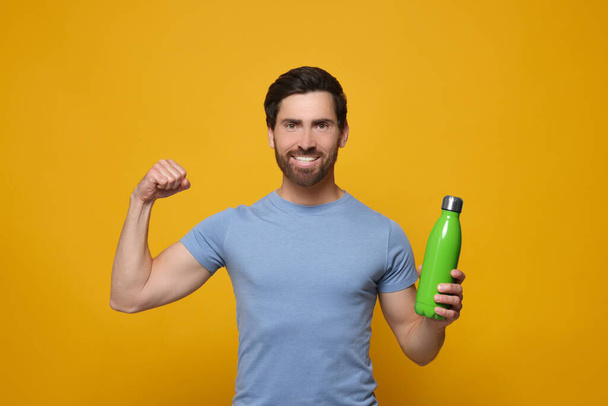 Man with green thermo bottle showing arm on orange background - Zdjęcie, obraz