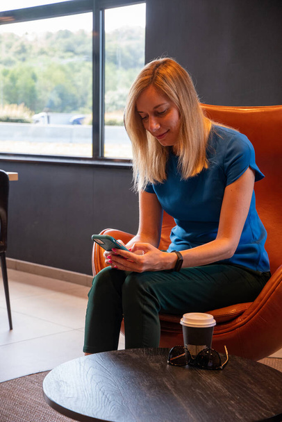 donna d'affari bionda positiva con in mano un telefono cellulare, felice di ricevere un messaggio dalla sua migliore amica o collega, seduta in un'accogliente caffetteria a tavola, foto di alta qualità - Foto, immagini