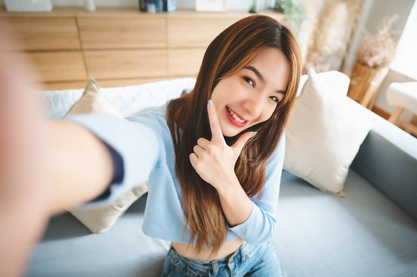 Csinos, fiatal ázsiai nő, nagy mosollyal a nappaliban. Élvezi, hogy vidám szelfit készít homályos háttérrel. - Fotó, kép