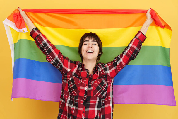 Persona androgena che alza una bandiera arcobaleno lgbt in studio con sfondo giallo - Foto, immagini