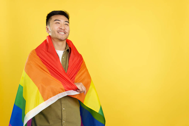 Happy chinese man wrapping with a lgbt rainbow flag in studio with yellow background - Photo, Image