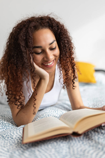 ベッドの上で本を読んでいるラテン系アメリカ人女性の垂直写真。余暇、文学、人々の概念 - 写真・画像