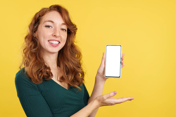 Redheaded ευτυχισμένη γυναίκα παρουσιάζει ένα κινητό με μια λευκή οθόνη στο στούντιο με κίτρινο φόντο - Φωτογραφία, εικόνα