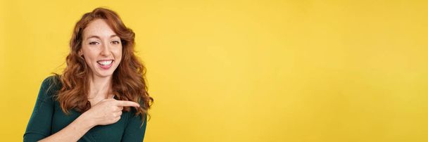 Redheaded woman pointing a blank space and smiling in studio with yellow background - Фото, изображение
