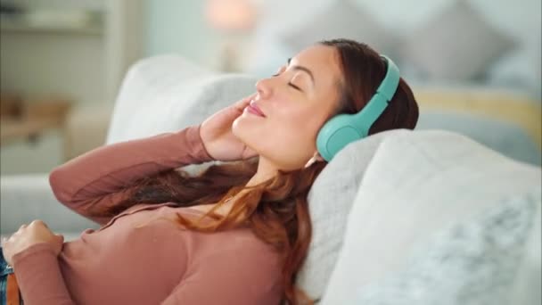 Słuchawki dla kobiet, słuchanie muzyki relaksacyjnej, podcast i uśmiech z spokojnym, zen i spokojny dzień w domu. Happy Asian kobieta i zrelaksowany kobieta siedzi na kanapie w salonie i medytacji z audio. - Materiał filmowy, wideo