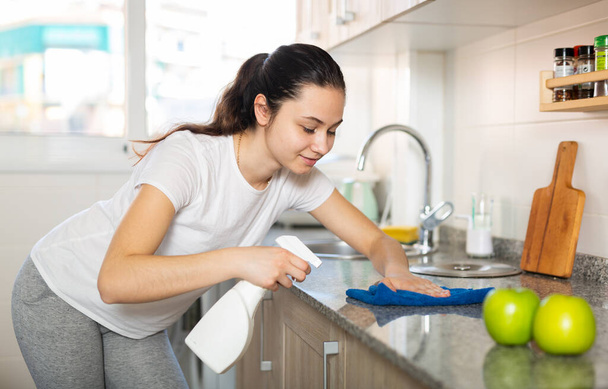 Young woman cleaning kitchen surfaces using sponge and spray cleaner - 写真・画像