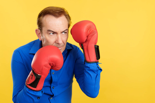 Un hombre maduro enojado con guantes de boxeo en estudio con fondo amarillo - Foto, Imagen