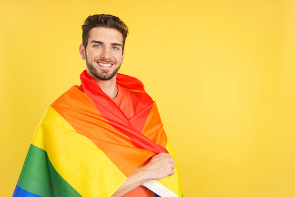 Happy caucasian man wrapped with a lgbt flag in studio with yellow background - Photo, Image