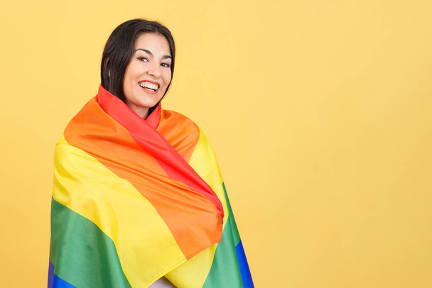Happy caucasian woman wrapped with a lgbt flag in studio with yellow background - Photo, Image