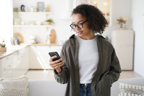 Focused young girl using smartphone at home, shopping groceries for kitchen in online store with mobile apps. Modern female in glasses chatting in social network, browsing content. - Foto, Bild