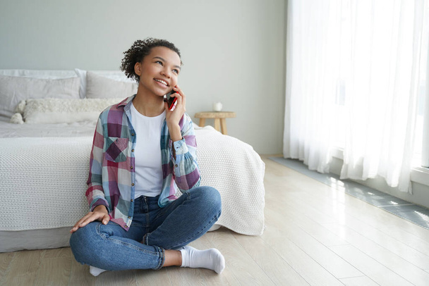 Ochtend telefoontje. Jonge Afro-Amerikaanse vrouw praat met een vriend op smartphone in de slaapkamer. Schattig spaans tienermeisje in casual kleding is telefoonverslaafde. Communicatie en ontspanning. - Foto, afbeelding
