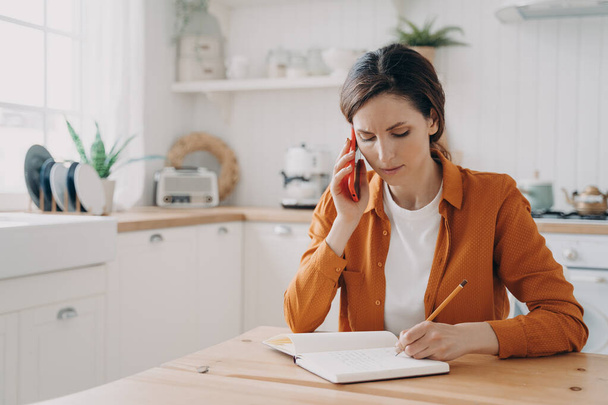 European girl is sitting at the kitchen, taking notes and talking on phone. Tired young woman is remote employee. Secretary, office manager or customer support assistant. Distance work concept. - Foto, Bild