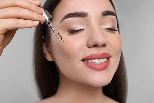 Happy young woman applying essential oil onto face on light grey background, closeup - Фото, зображення