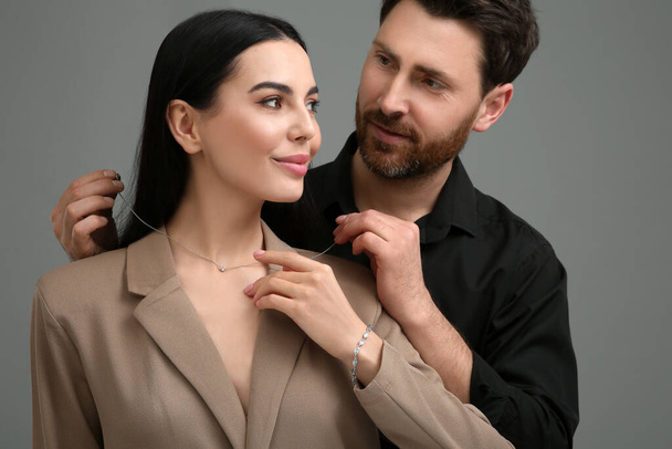 Man putting elegant necklace on beautiful woman against dark grey background - Valokuva, kuva