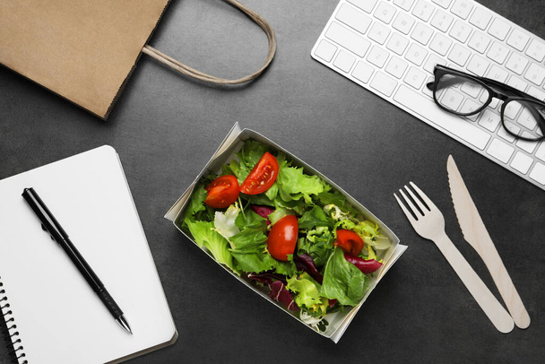 Container of tasty food, keyboard, cutlery and notebook on grey table, flat lay. Business lunch - Фото, изображение