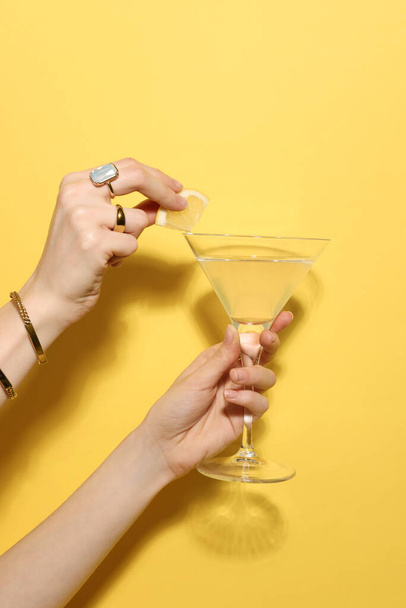 Woman adding lemon slice to martini glass of refreshing cocktail on yellow background, closeup - Foto, afbeelding