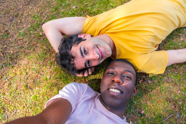 Couple of multi-ethnic men in a park, lgbt concept, lying on the grass taking a selfie - 写真・画像
