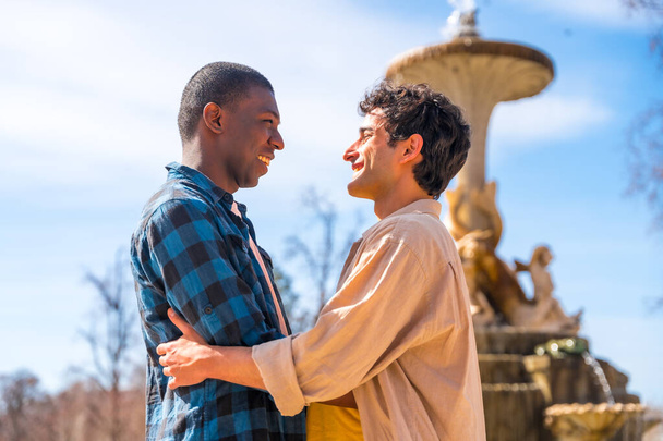 Couple of multiethnic men in a city fountain, lgbt concept, in a romantic pose looking at each other smiling - Photo, Image