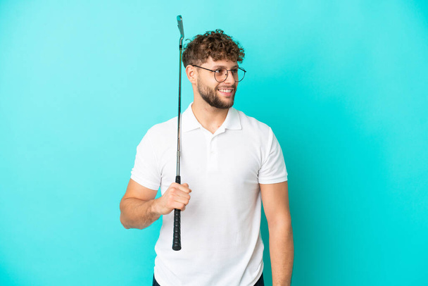 Young handsome caucasian man isolated on blue background playing golf - Fotoğraf, Görsel