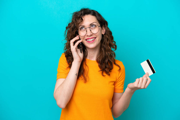 Young caucasian woman isolated on blue background keeping a conversation with the mobile phone and holding a credit card - Foto, afbeelding