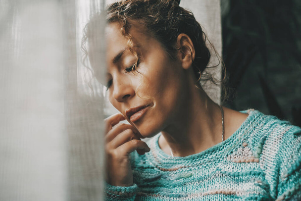 Exhausted and sad woman alone at home near the window with closed eyes and depressed expression on face. Closeup portrait of adult female people with sadness. People and mental burnout problems life - Photo, Image