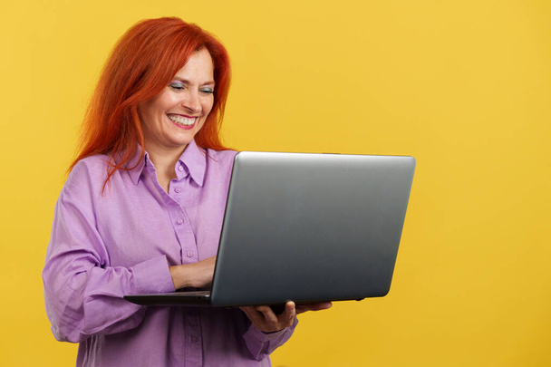 Piros hajú érett nő mosolyog használata közben egy laptop stúdióban sárga háttér - Fotó, kép