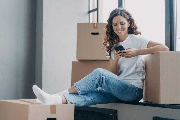 Happy spanish woman is sitting on windowsill of new home. Girl is unpacking boxes. Young lady is texting on phone and smiling. Joy of relocation concept. - Foto, afbeelding