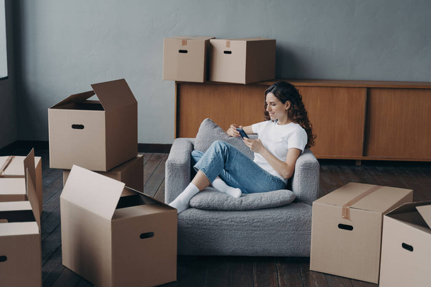 Woman in armchair clicks phone among boxes. European girl having rest in new apartment after relocation. Successful independent woman is moving. Mortgage loan and dream house purchase concept. - Фото, изображение