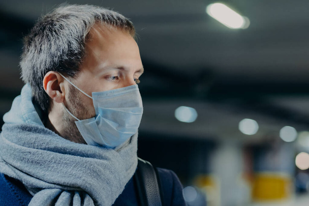 Pensive man wears protective mask against new coronavirus from China, wrapped scarf around neck, looks somewhere, thinks about epidemic situation. Influenza, flu symptoms, virus, treatment concept - Foto, imagen