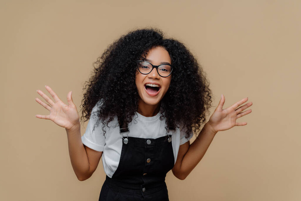 Overjoyed African American woman laughs out, raises palms, being in high spirit, wears transparent glasses, casual t shirt and overalls, poses against brown background, feels happiness and pleasure - Photo, Image