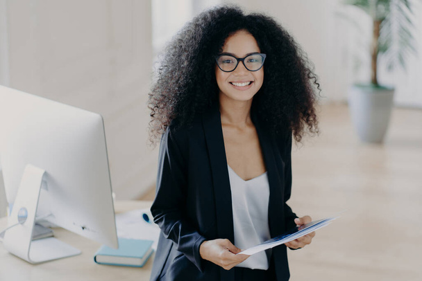 Photo of lovely African American woman holds some papers, being consultant manager, wears elegant clothes and spectacles, stands near desktop with computer and textbooks. Happy businesswoman - Fotó, kép
