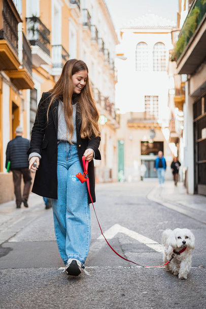 Wesoła młoda kobieta spacerująca obok swojego białego psa w słonecznym mieście - Zdjęcie, obraz