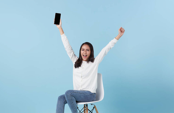 Feliz sorrindo mulher asiática segurando smartphone sentado na cadeira e ganhando o prêmio no fundo do estúdio azul. - Foto, Imagem
