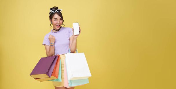 Feiern asiatische Frau lächelt beim Einkaufen, zeigt sie Smartphone-App Online-Shopping auf gelbem Hintergrund mit Einkaufstaschen. Volle Länge  - Foto, Bild