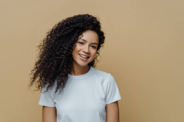 Portrait of beautiful woman with frizzy bushy hair, gentle smile and healthy skin, wears casual white t shirt, enjoys pleasant conversation stands against beige background copy space. People, emotions - Foto, afbeelding