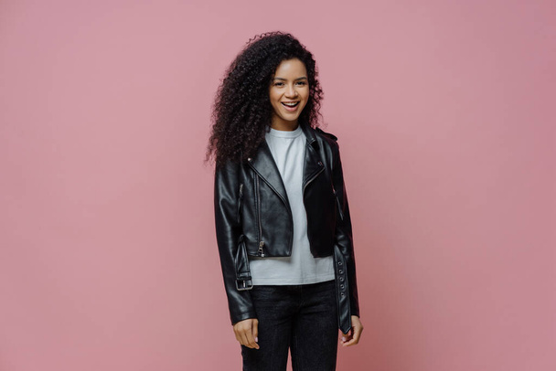 Happy positive Afro American woman dressed in leather jacket, ready for outdoor walk, poses against pink background. Joyful dark skinned millennial girl in good mood has casual talk with friend - Φωτογραφία, εικόνα