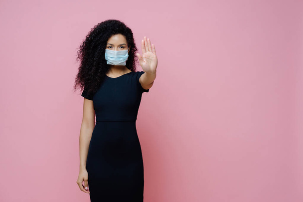 Epidemia Covid 19, choroba wirusowa. Zdjęcie etnicznej kobiety z kręconymi włosami sprawia, że stop gest z dłoni, mówi nie koronawirusowi, nosi ochronną sterylną maskę, aby uniknąć wirusa, ubrany w czarną sukienkę - Zdjęcie, obraz