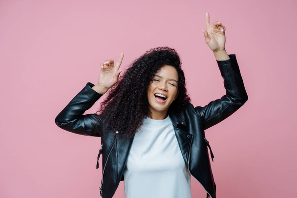 Carefree beautiful Afro American woman dances with arms raised, moves with rhythm of music on disco party, wears white t shirt and leather jacket, closes eyes with pleasure, keeps mouth opened - Foto, afbeelding